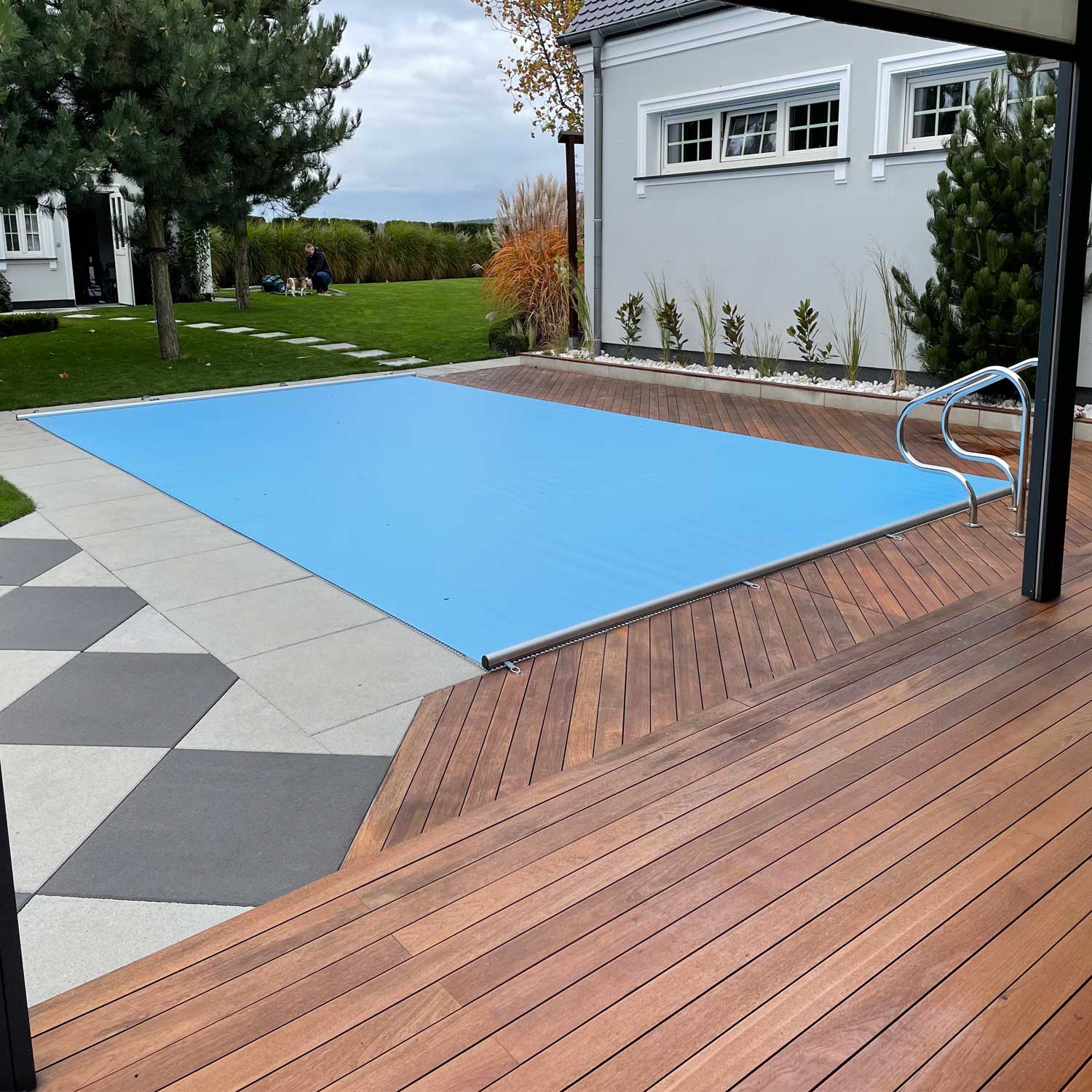 Der rechteckige Pool mit blauer Sicherheitsabdeckung liegt auf einer Holzterrasse und Steinfliesen.
