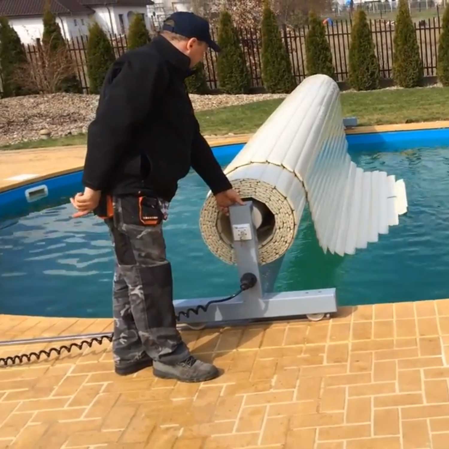 Mann bedient ein angetriebenes Rollensystem zum Aufrollen einer weißen Schwimmbadabdeckung mit Lamellen.