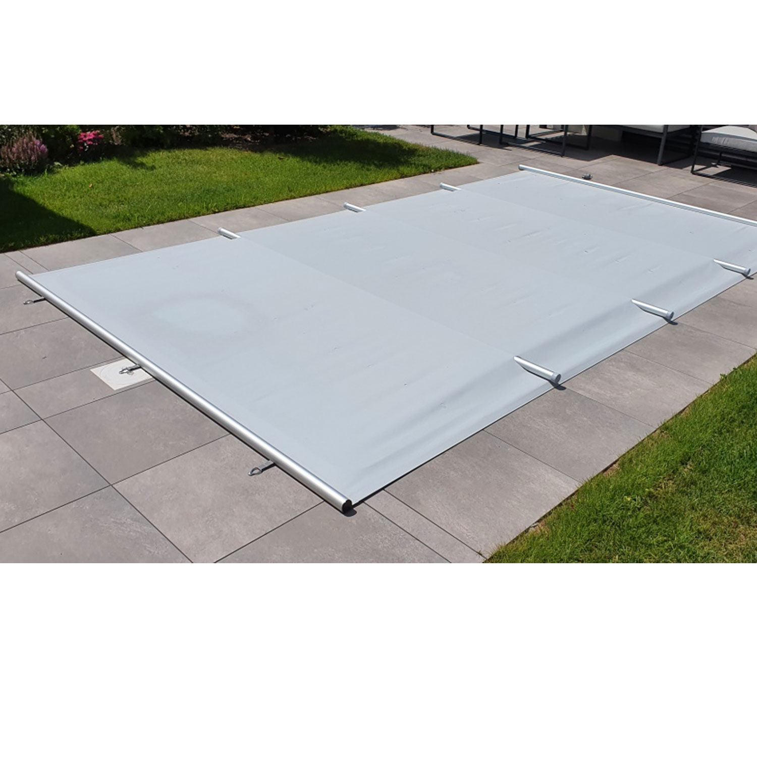 Rechteckiger Pool mit grauer Sicherheitsabdeckung abgedeckt und mit Metallstangen gesichert auf einer grau gefliesten Terrasse.